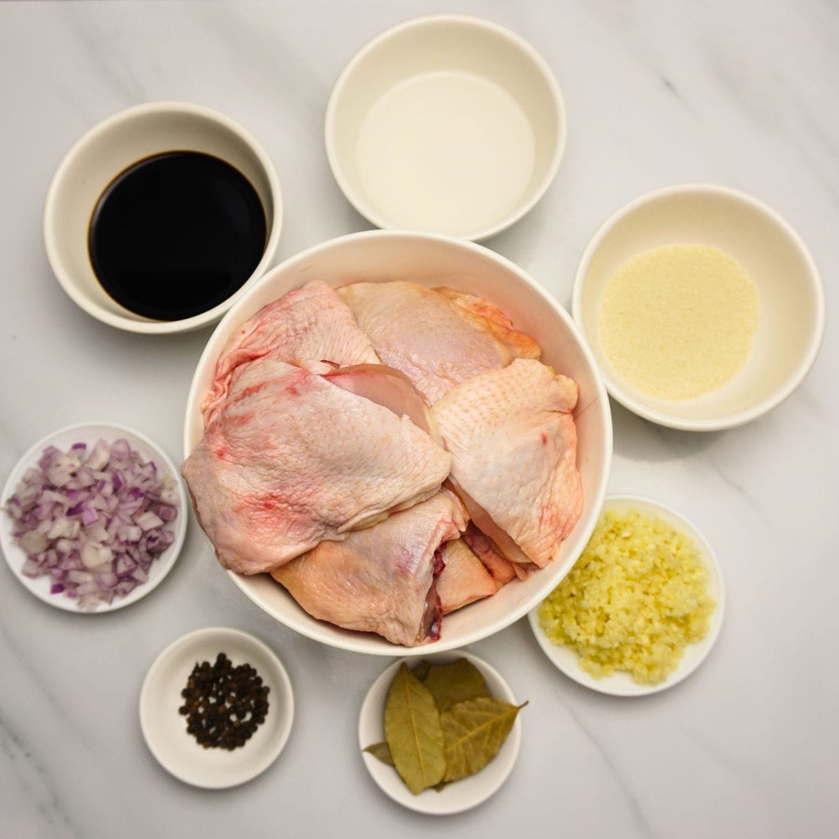 Chicken thighs, soy sauce, vinegar, oil, garlic, laurel, whole black pepper and onions in ceramic bowls.