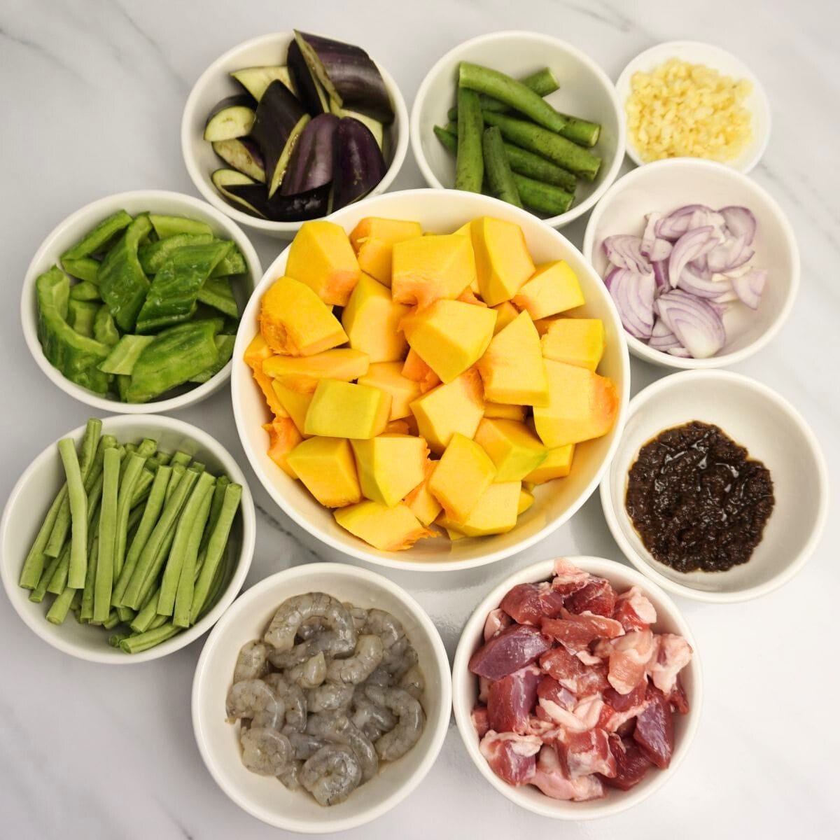 Squash, okra, eggplant, bitter gourd, string beans, garlic, onion, shrimp paste, shrimp and pork in bowls.