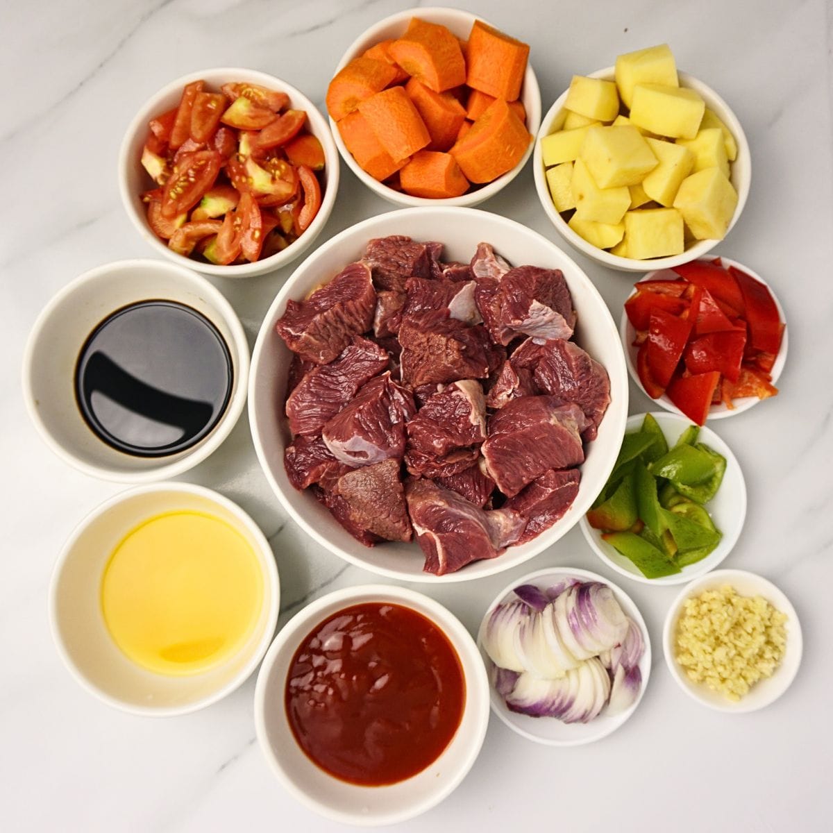 Beef mechado ingredients in bowls. Beef brisket, tomato sauce, soy sauce, bell peppers, carrots, potatoes, tomatoes, onion and garlic.