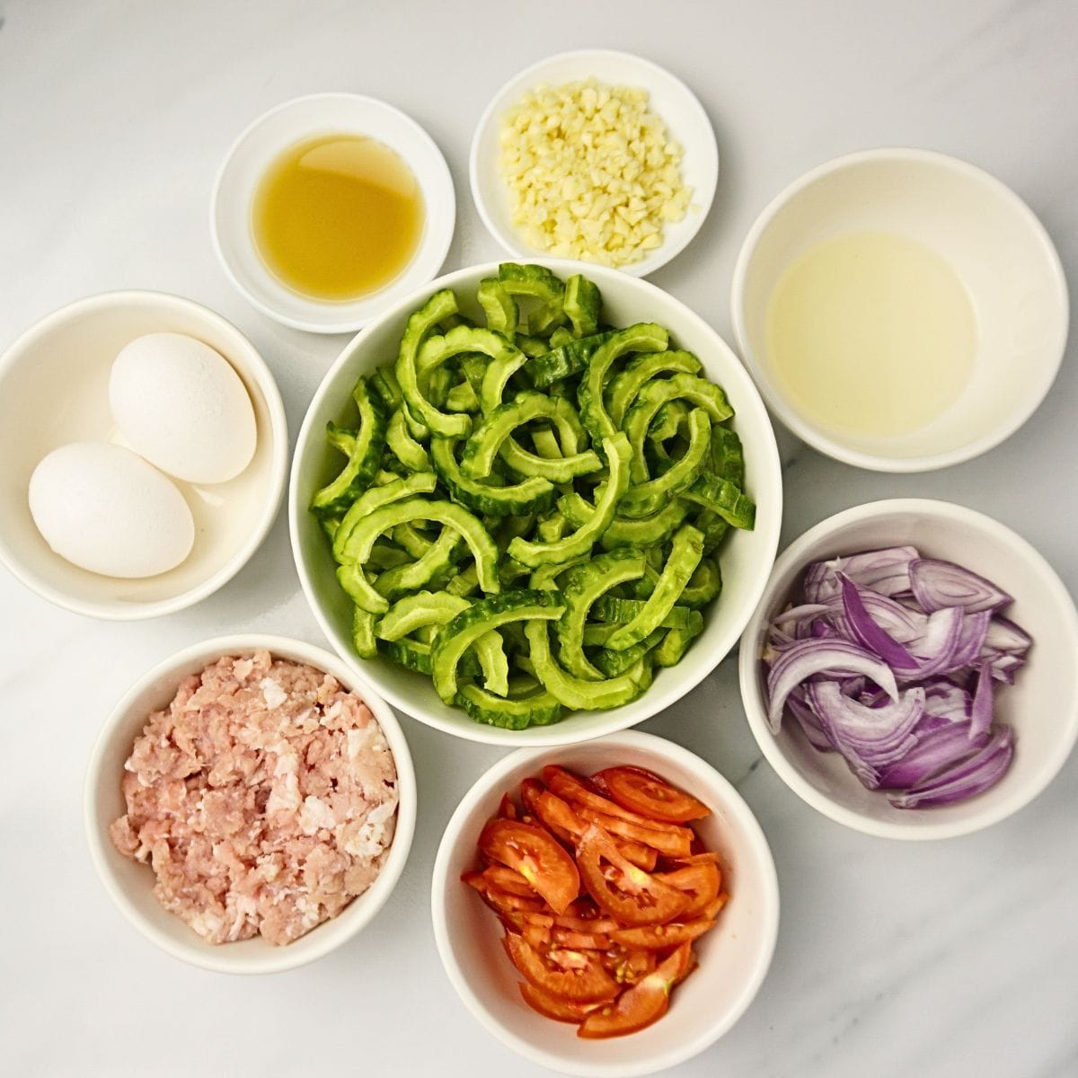 Ginisang ampalaya ingredients in bowls. Bitter gourd, ground pork, egg, fish sauce, garlic, oil, onion and tomatoes.
