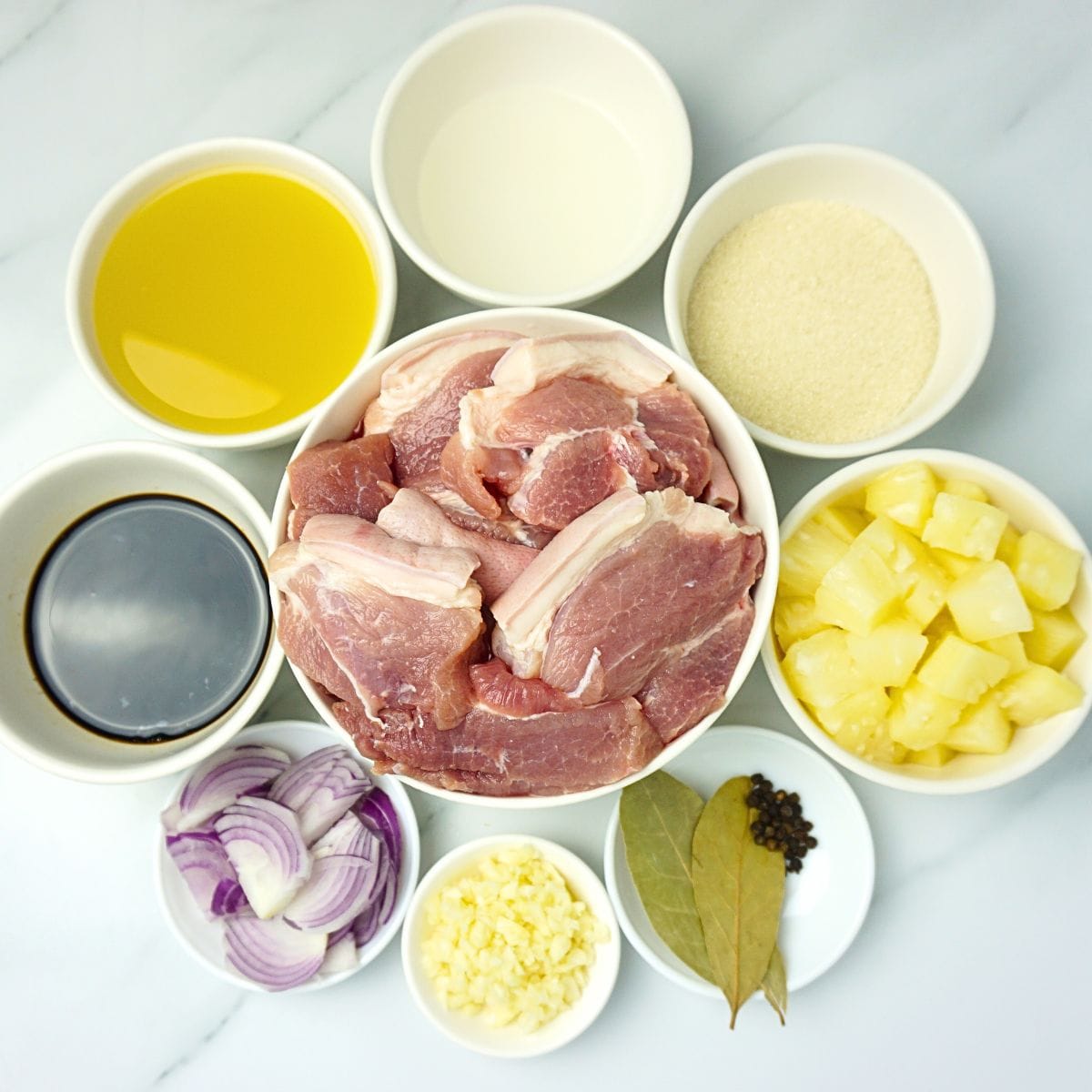 Pork hamonado ingredients in bowls. Pork shoulder, garlic, onions, soy sauce, pineapple juice, vinegar, sugar, pineapple chunks, bay leaves and black pepper.