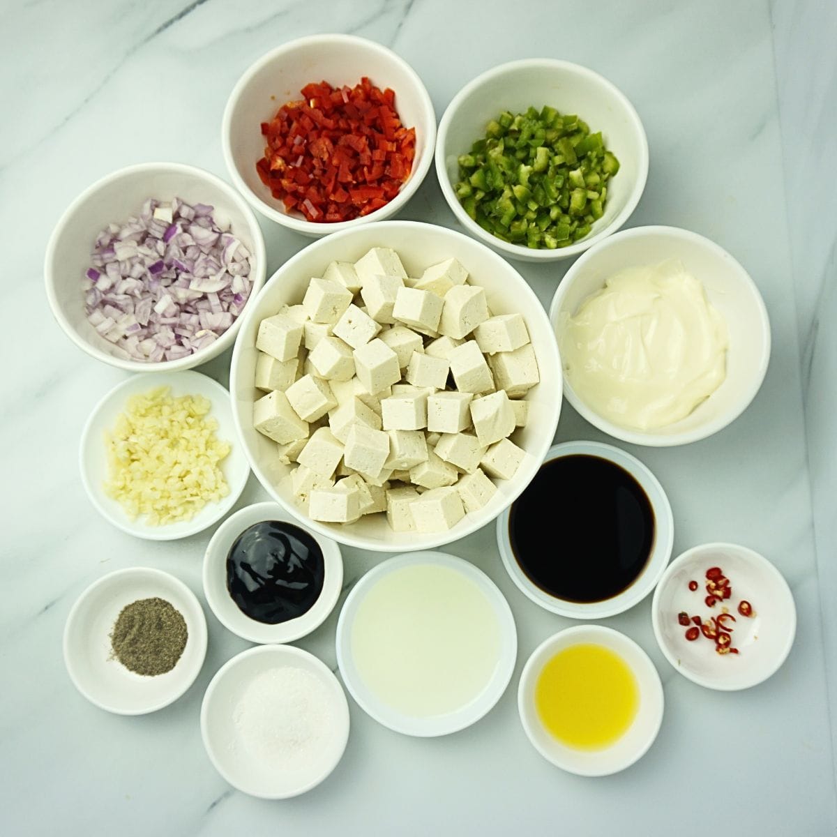 Tofu sisig ingredients in bowls. Tofu, garlic, onion, red bell pepper, green bell pepper, sugar, soy sauce, oyster sauce, chili, calamansi juice, black pepper and mayonnaise.