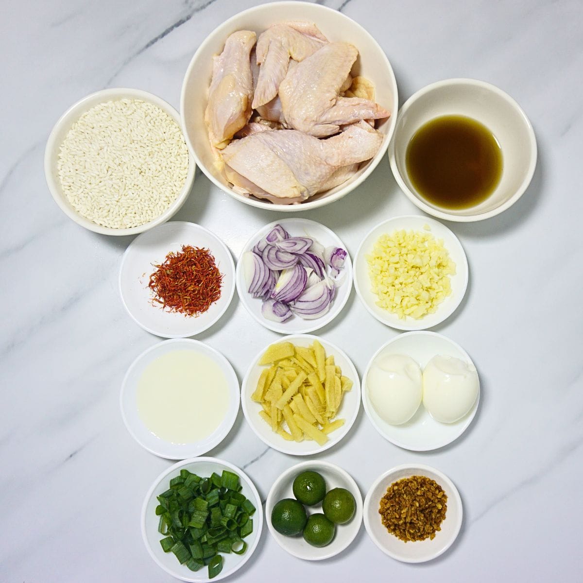 Arroz caldo ingredients in bowls. Glutinous rice, sliced chicken, fish sauce, kasubha, onions, garlic, cooking oil, ginger, boiled eggs, green onions, calamansi and fried garlic.