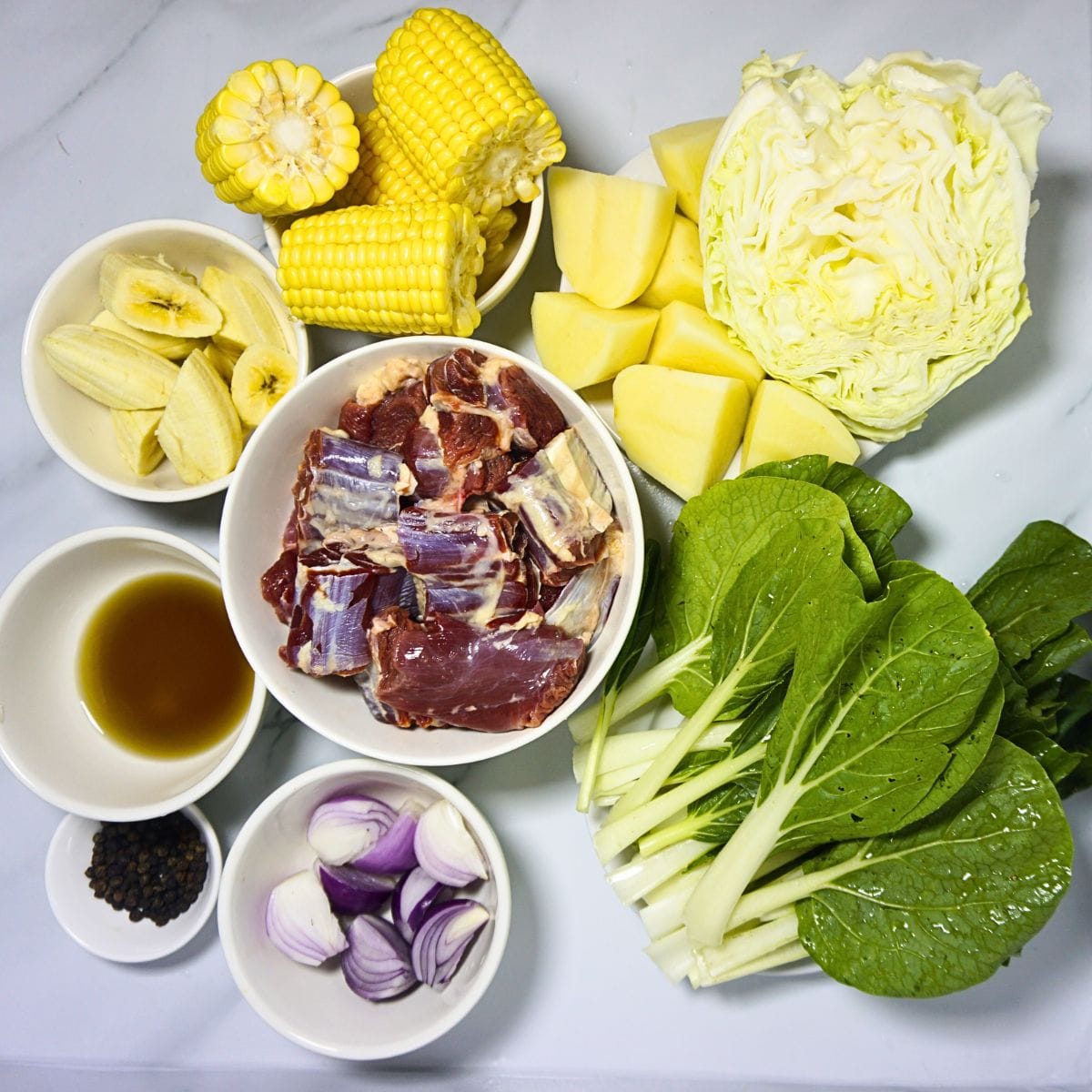 Nilagang baka ingredients in bowls. Beef shanks, onion, whole pepper, fish sauce, sweet plantain, sweet corn, potatoes, cabbage and pechay.