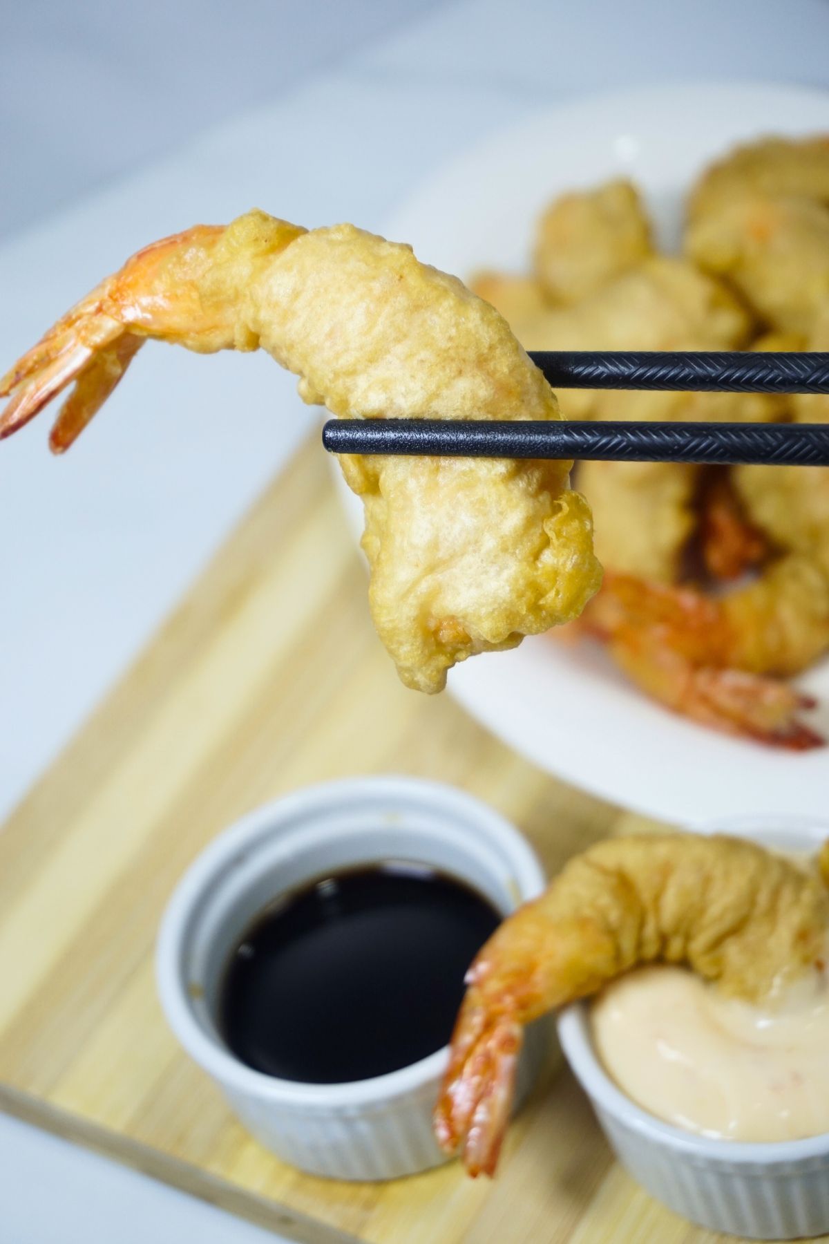 Chopsticks holding a tempura with dipping sauce on a wooden cutting board.