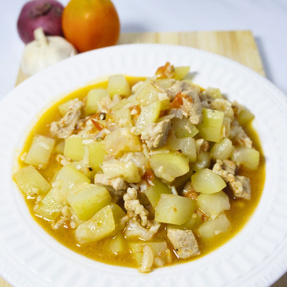 Sauteed bottle gourd in a plate with fresh garlic, onion and tomato on a wooden cutting board.