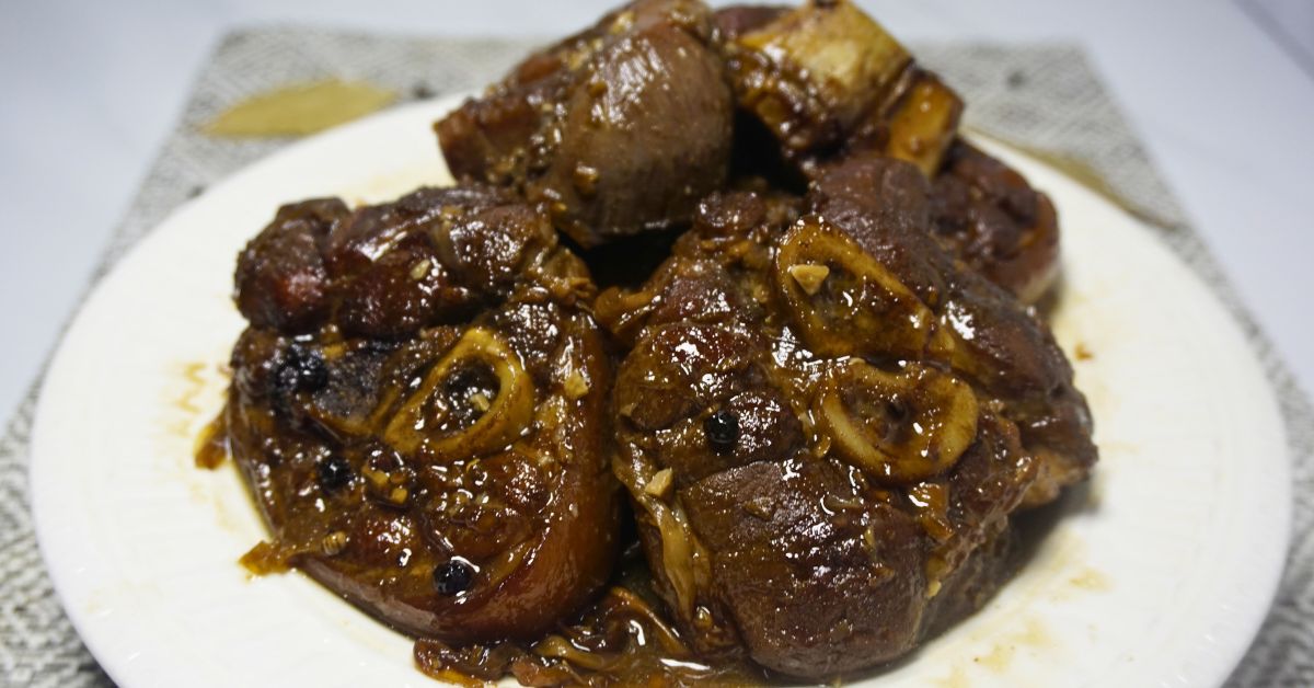 Paksiw na pata in a plate with dried bay leaves and peppercorns on a table.