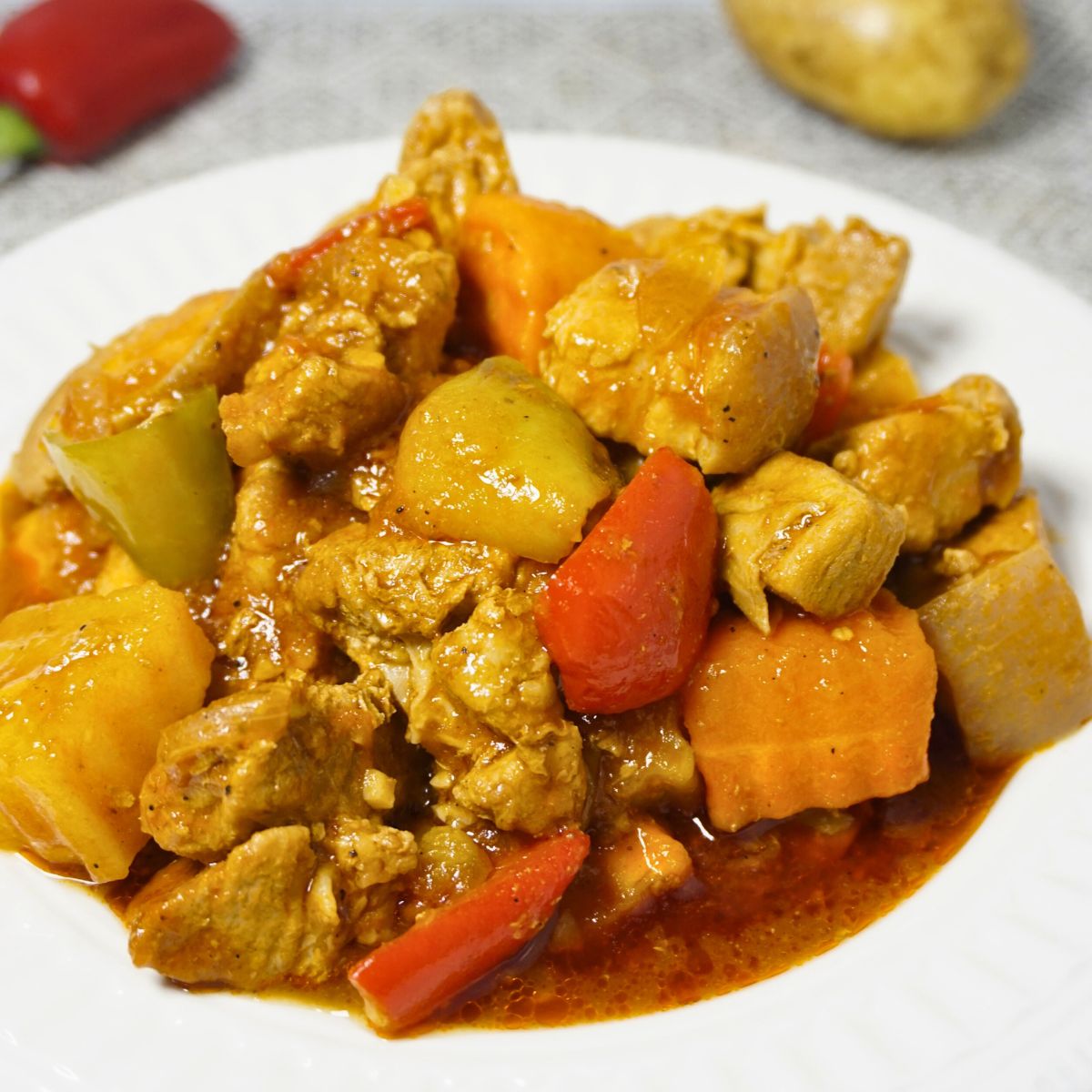 Plate of pork afritada with raw potato and bell pepper on a table.