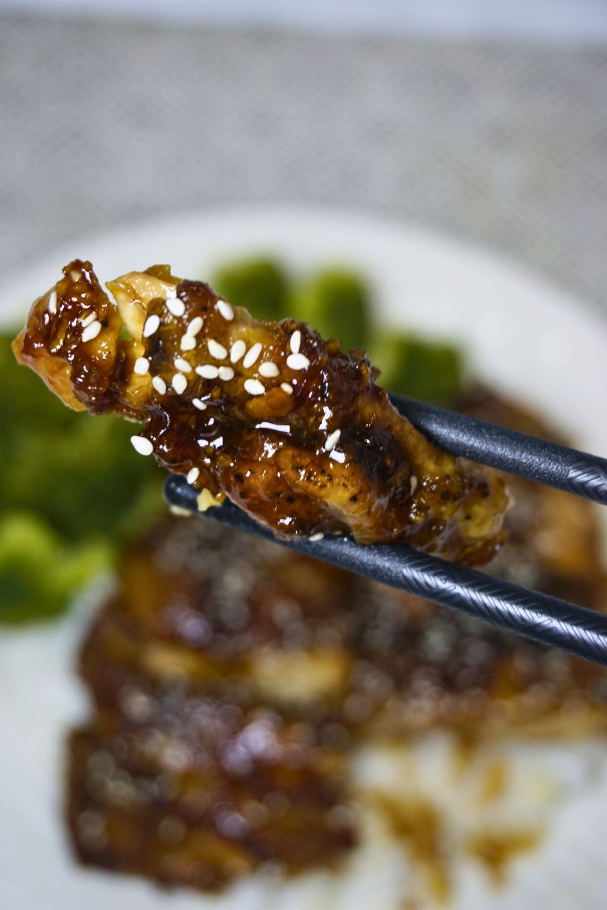 Chopsticks holding a piece of chicken teriyaki with sesame seeds.