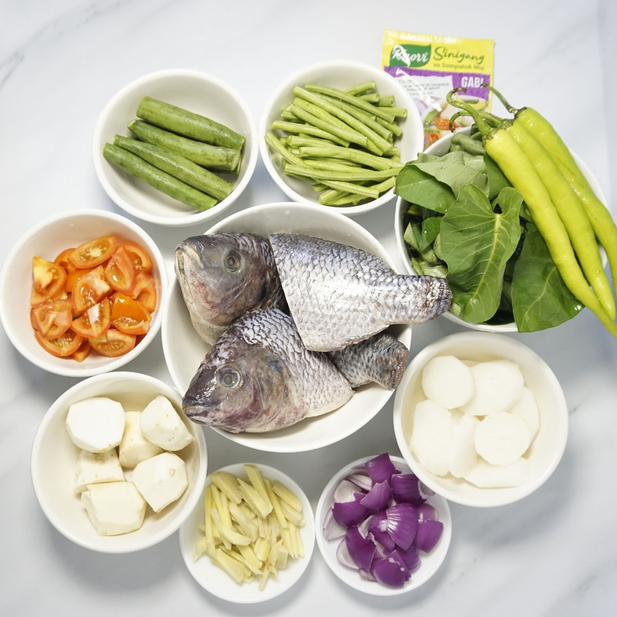 Sinigang ingredients in bowls. Tilapia, onion, ginger, radish, tomatoes, okra, snake beans, kangkong and green chilies.